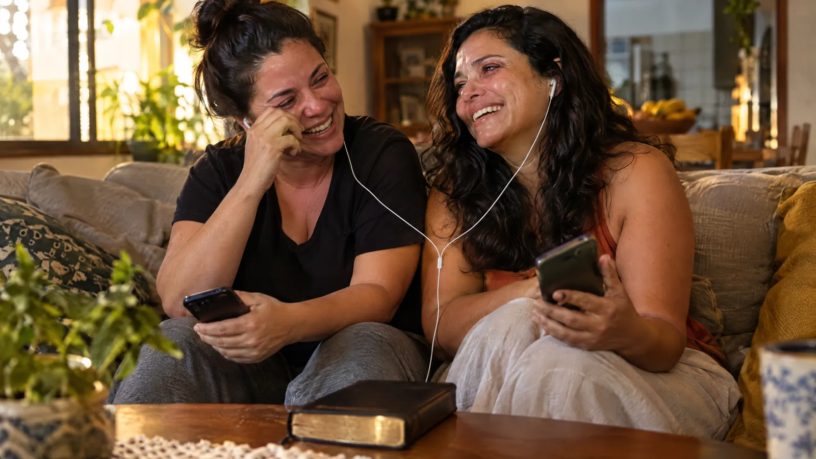 Duas amigas evangélicas dividindo o fone e rindo emocionadas no sofá